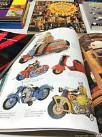 Open pages showing vintage tin toy motorcycles and scooters plus several closed books in background