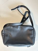 Front view of rectangular black Monos shoulder bag, showing logo, top zipper and strap