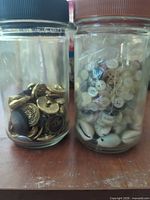 Two glass jars; left filled with gold and dark metal buttons, right with small white buttons and cowrie shells