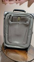 Front view of gray Travelpro carry-on suitcase showing zippered front pocket and foot stands