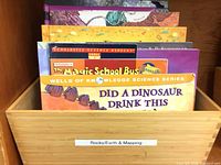 Books in wooden bin labeled Rocks/Earth & Mapping, visible Magic School Bus and Did a Dinosaur Drink This Water?