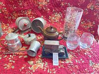 Group shot showing all items including teapot, aluminum cookware, glass candle holder, two glasses, Banthrico car bank on tray, tablecloth background
