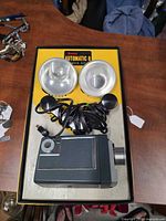 Box interior showing two flash reflectors, coiled power cord, and side view of camera