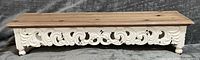 Front view of wood riser showing natural top, carved white apron, bun feet