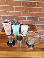 All items arranged on table: two travel tumblers, ceramic mug, gravity steeper, two infusers, straw, brush