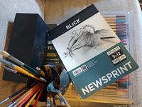 Arteza paint set box, Blick newsprint pad, colored pencil case, cup of assorted brushes