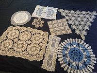 Group view of nine crochet and lace pieces laid out on dark cloth