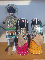 Group shot of three Ndebele beaded dolls showing relative sizes, colours, and attached tags.