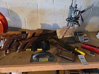 Wide view of tabletop with hand saws, mitre box, float, stapler, safety goggles, drill press column