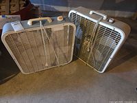 Two white box fans standing side by side on concrete floor