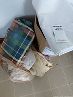 Stack of loose fabric pieces including plaid panel, beige knit throw, off-white fabric and additional piece in bags