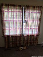 Pair of tan and burgundy plaid curtain panels on rod with rolled matching bed skirt below window