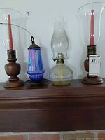 All four items lined on mantle: two wooden hurricane candle holders with glass chimneys and red candles, one iridescent hanging lantern, one clear glass kerosene lamp with chimney