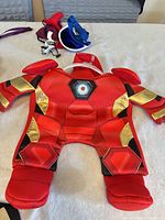 Front view Iron Man dog costume with assorted hats, bow tie, devil horns in background