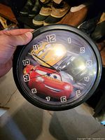 Front view of round black-rim wall clock with Lightning McQueen and yellow Dinoco car graphic, visible numerals and hands
