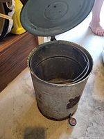 Overall view of cylindrical metal trash can with lid open, showing interior liner and foot pedal