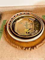 Stack of assorted trays showing gold bird tray on top, black patterned rim tray beneath, rope-edge tray at bottom