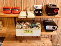 Shelf with two red viewers, black viewer, reel album, box of picture packets, two projectors