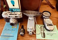 Front view of Pax M4 camera, lens, instruction booklet, Sunpak adapter, flash gun, Sekonic meter, leather case and GE flashbulb box visible