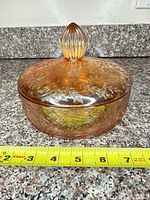 Side view of amber carnival glass covered dish with lid showing overall form and floral pattern, measuring tape indicates size