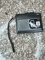 Front view showing Optimus CTR-115 cassette recorder with speaker grille, VOX label and attached wrist strap