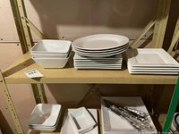 Stacks of square bowls, oval plates, square plates, and wrapped knives on shelves