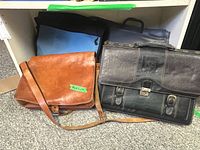 Front view of brown leather messenger and black KS Design briefcase, other two bags partially visible behind