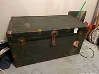 Front view of green metal trunk showing brass latches, center lock, travel tag