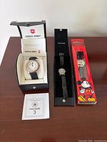 All three watches displayed: Swiss Army in open case with manual, Movado in black box, Disney Mickey watch sealed in red retail box