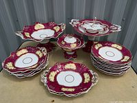 Group shot showing entire wine red and gold porcelain dinnerware set including stacks of plates, serving platters, compote and pedestal stand