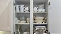 Overview of cabinet showing stacks of cream dinnerware, clear glass plates, and white mugs