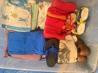 Plastic bin with folded toddler clothes, red rain boots, blue pants, plaid shirt, flip-flops, brown shoes, beige hoodie, yellow-blue sneakers