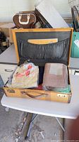 Open medium tan woven suitcase with leather trim showing interior contents – blankets and clothing
