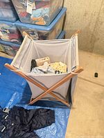 Wood-frame mesh hamper containing baby clothing; snow bib pants visible