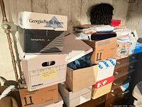 Stacked cardboard boxes with papers, wig on top, ribbon spools visible
