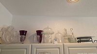 All items on cabinet top: glass cake stand with dome, additional cloche, purple vases, tumblers, stacks of bowls and plates, metal stand with square plates