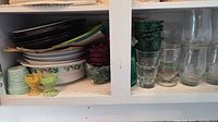 Cabinet shelf showing stacked plates, bowls, casserole dishes, green ruffled bowls, pastel egg cups, clear etched tumblers and small glasses