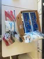 Cardboard box holding three Kirkland 360-piece cutlery boxes, Hefty storage bags pack, and multiple bags of assorted colored plastic cutlery
