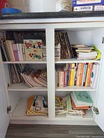 Cabinet shelves filled with assorted vintage cookbooks, pamphlets, spiral notebooks and recipe card box