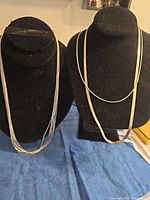 Two silver-tone necklaces displayed on black necklace forms, left shows multi-strand beaded design, right shows flat herringbone chain