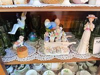 Shelf showing lady figurine, dog figurine, floral wheelbarrow, decorative eggs, art glass egg, green glass tumblers, and small wood box