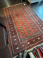 Full view of rust red Bokhara rug on floor showing borders and fringe