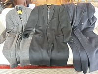Three overcoats laid side by side (grey herringbone, black, navy) showing front view and overall condition