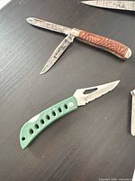 Green synthetic handle folding knife with drilled handle and partially serrated blade next to brown jigged bone two-blade knife