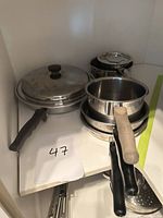 Stacked view of cookware pieces inside cabinet showing saucepans, large lidded pan, handles, steamer insert