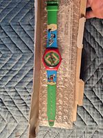 Full view of wrist watch in open box showing green strap, blue character panels, red bezel, square dial