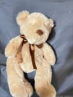 Full view of beige plush teddy bear with brown ribbon lying on fabric