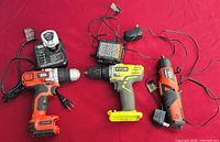 Three cordless power tools with chargers laid out on red surface