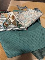 Stack of three fabric remnants showing teal geometric, pastel floral, and scenic stripe pieces