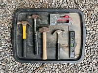 Tray displaying four hammers, staple gun, and torpedo level on gravel